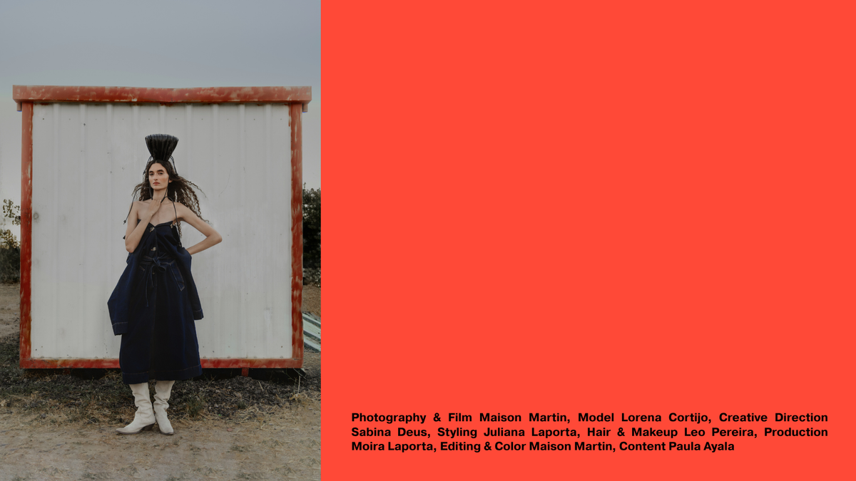A fashion editorial-style photo with a model wearing a dark navy dress and white boots standing in front of a white box with a red frame on the left, and a large solid coral-red block occupying the right side. Small text at the bottom right lists photography and production credits.